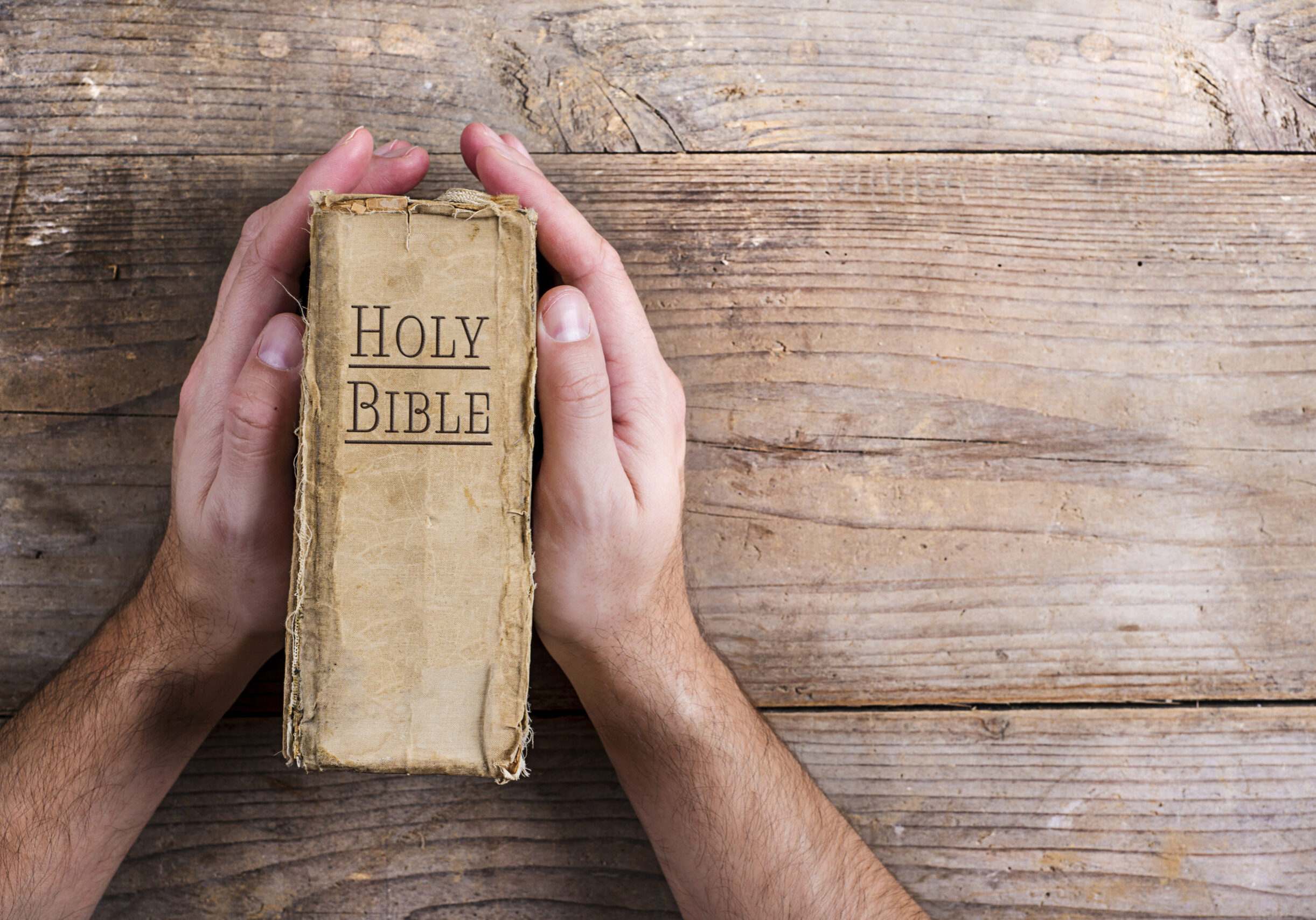 Hands holding Bible on a wooden desk background.